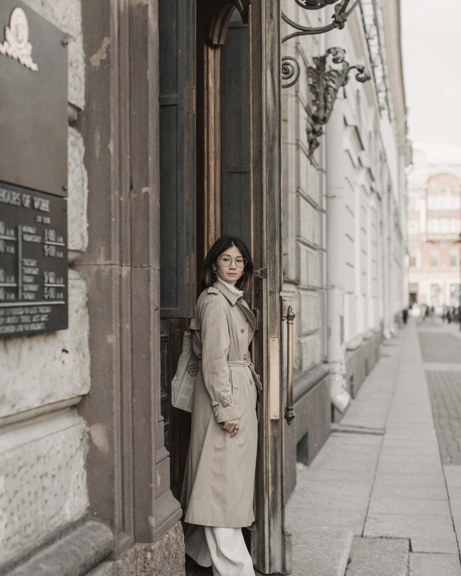 A woman in a stone cotton trench coat stands in the tall wooden doorway of a stone building on a city street